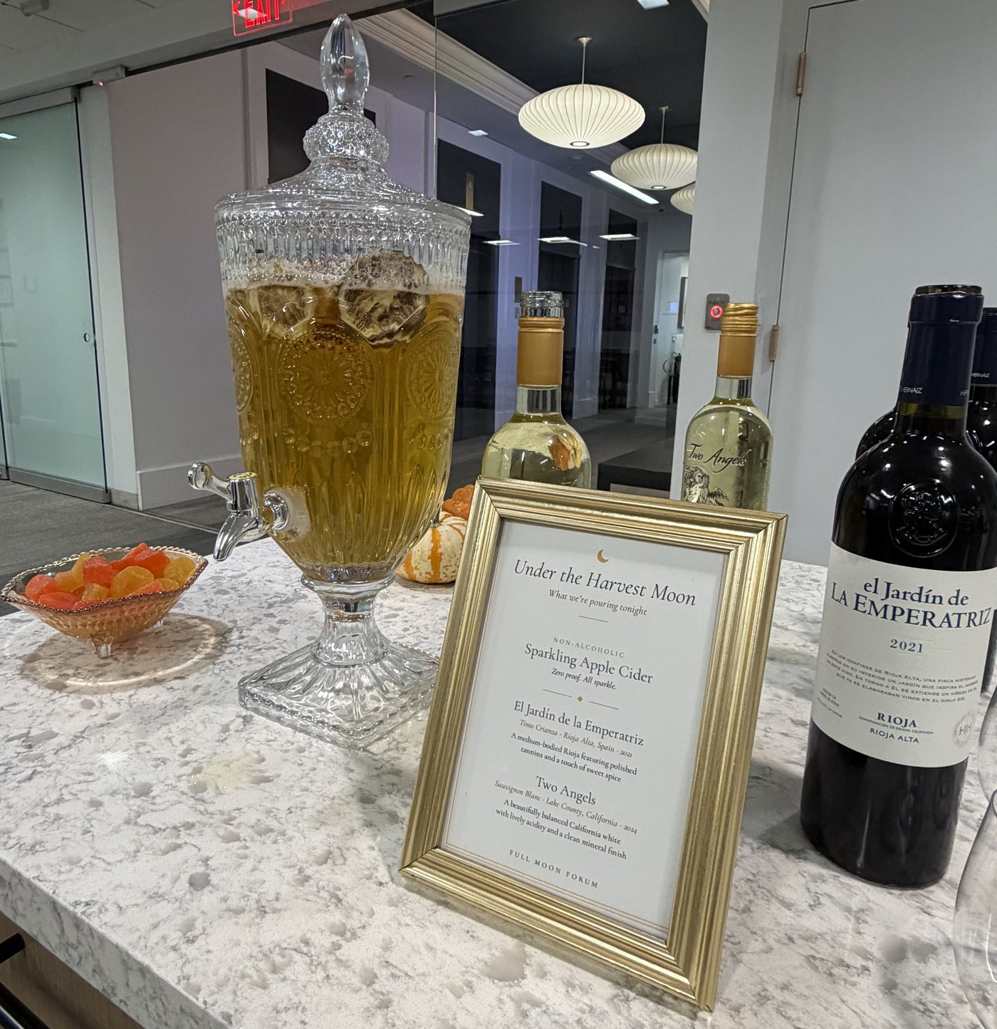 A glass dispenser of sparkling apple cider beside wine bottles and a gold-framed Under the Harvest Moon drinks menu
